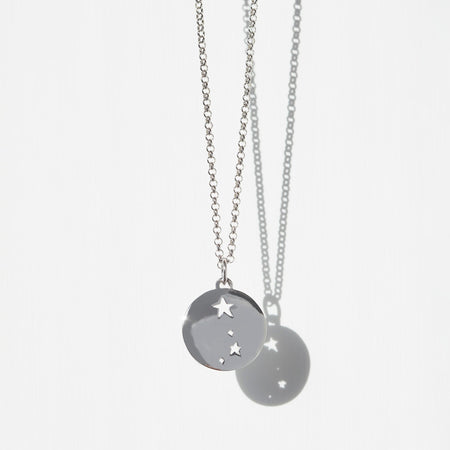 silver aries constellation disc charm on a silver belcher chain necklace in front of white background with shadow.